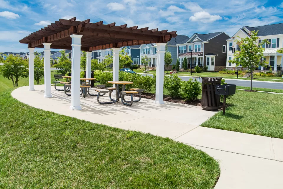 A pergola with a grill is located in a grassy park, across the street from single-family housing in a suburban style.